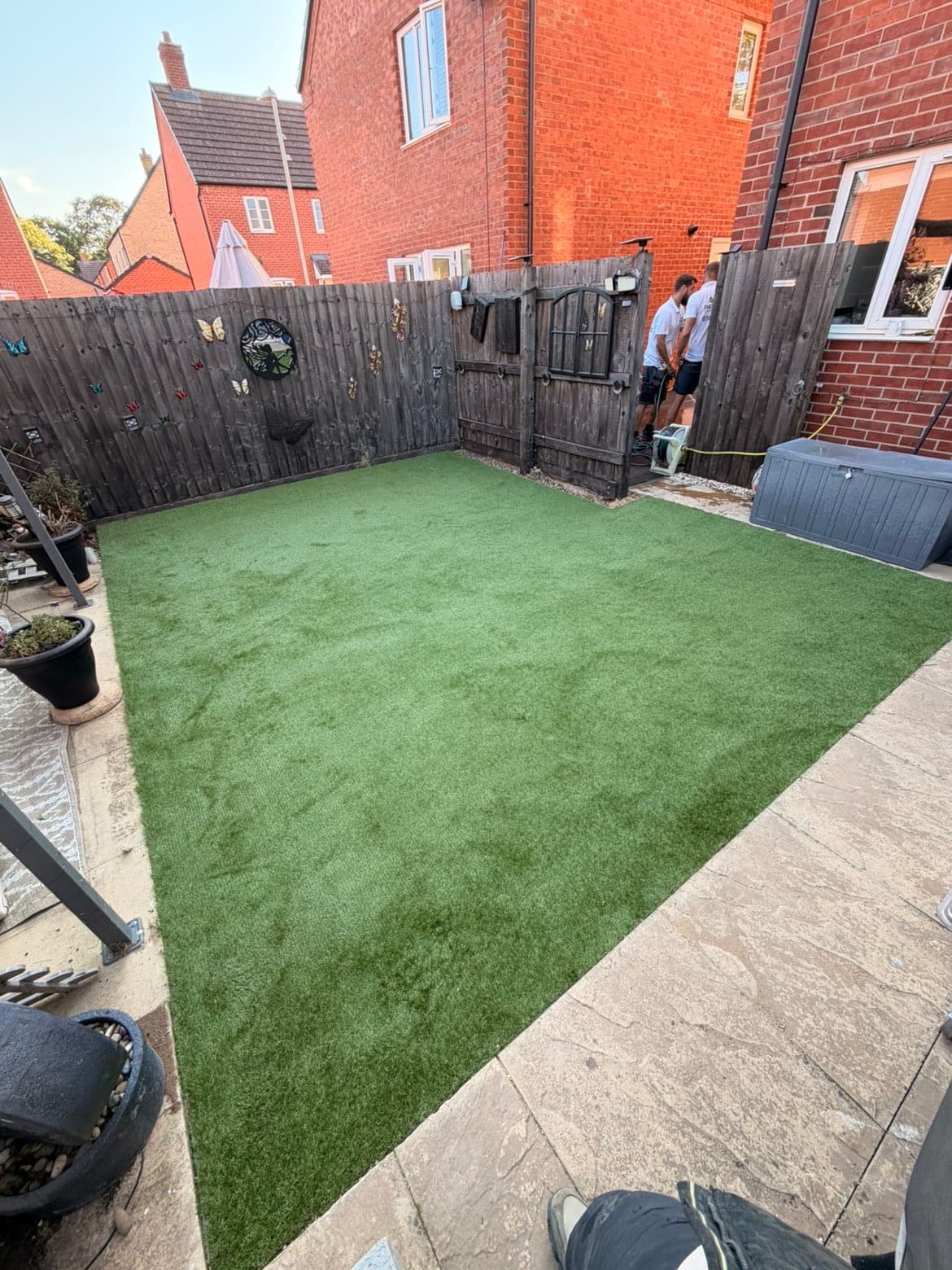 Completed artificial grass lawn cut cleanly along a diagonal edge against sandstone paving, with closeboard fencing and the house behind