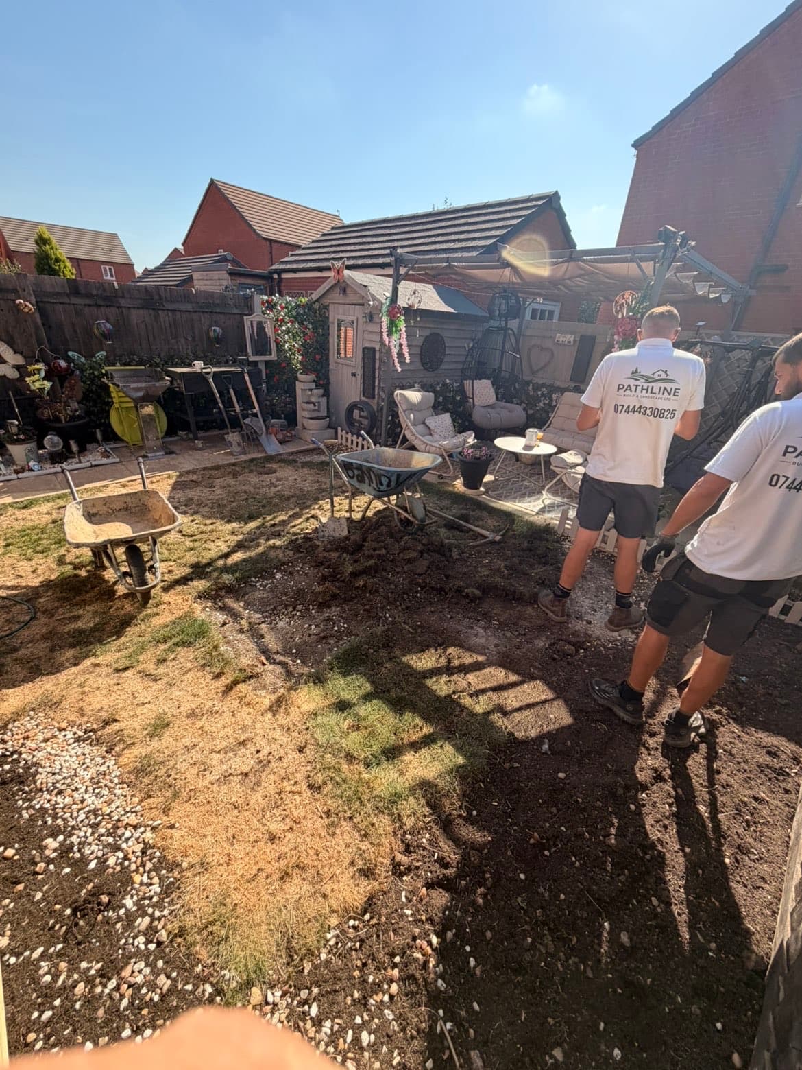 Before photo of a scorched patchy lawn being stripped out by two Pathline team members in branded shirts, with wheelbarrows and spades on site