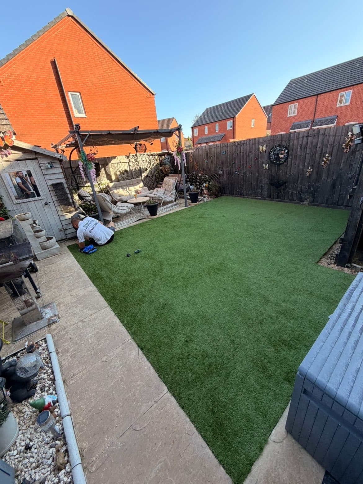 Pathline team member kneeling to trim and finish the edge of a newly laid artificial grass lawn beside the pergola seating area