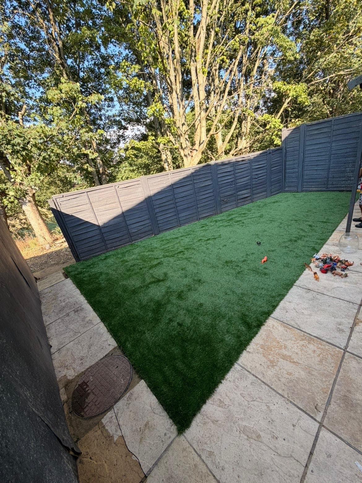 Completed full-width artificial grass lawn finishing flush against pale paving, with a dark grey boundary fence and mature trees behind