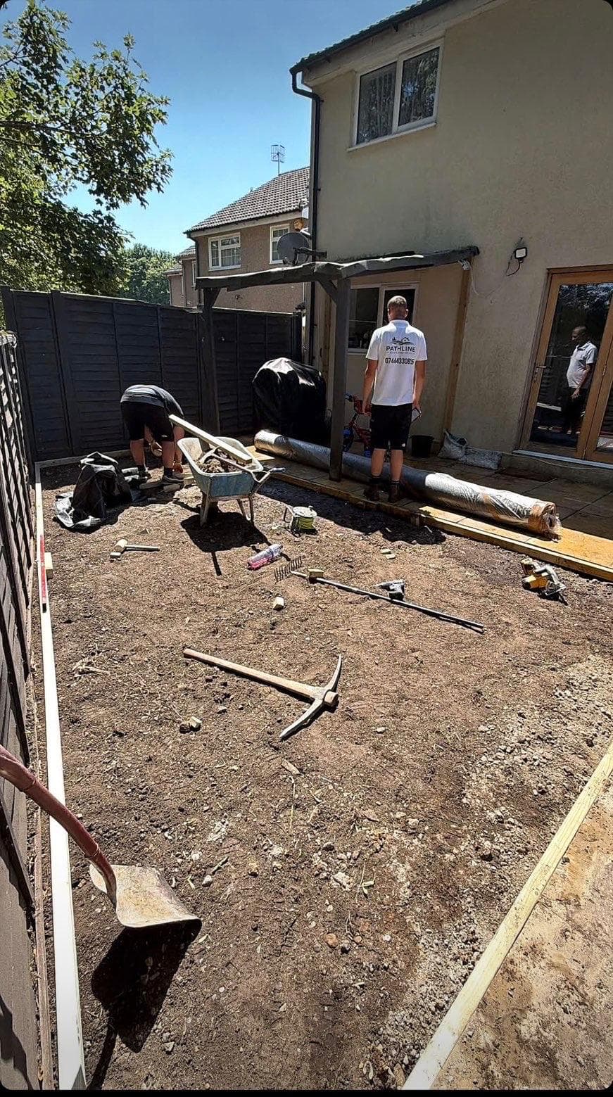 Rear garden dug out to bare soil with timber battens set around the perimeter, Pathline team members on site with a wheelbarrow and tools, and rolls of artificial grass and underlay ready against the wall