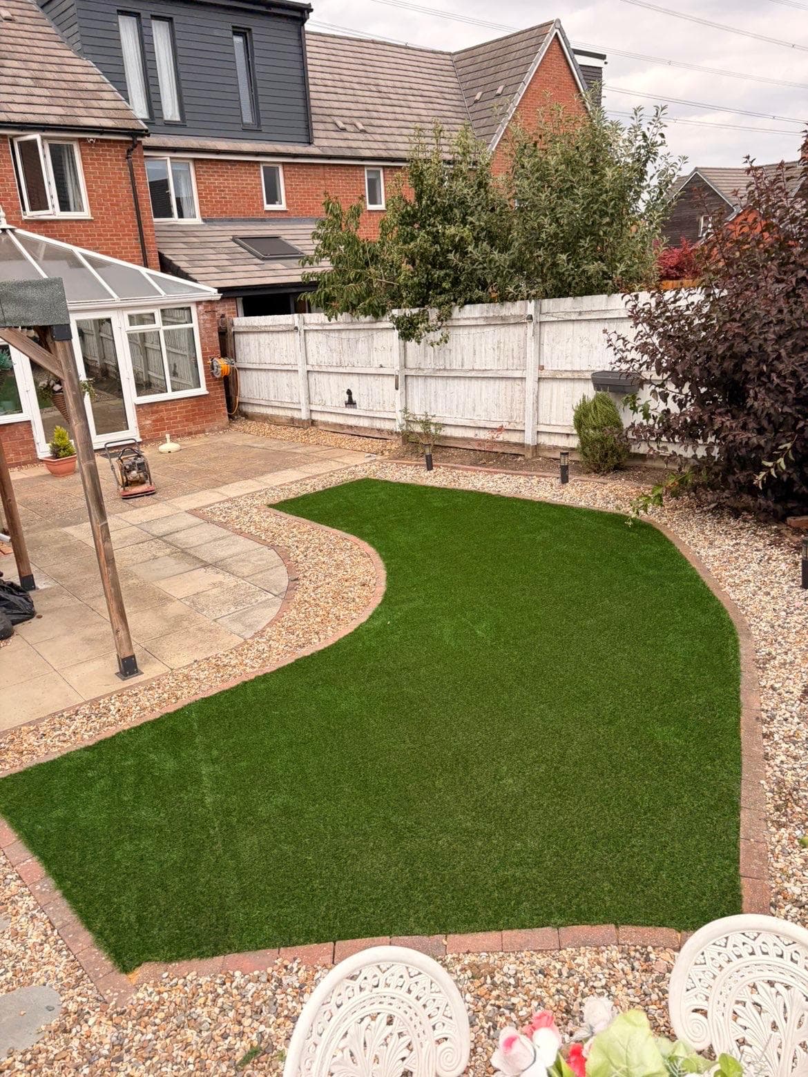 Completed curved artificial grass lawn with brick edging and pale gravel borders, beside a sandstone patio and pergola